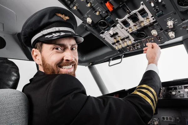 Cheerful pilot in cap reaching overhead panel with set of switches in airplane simulator — Stock Photo