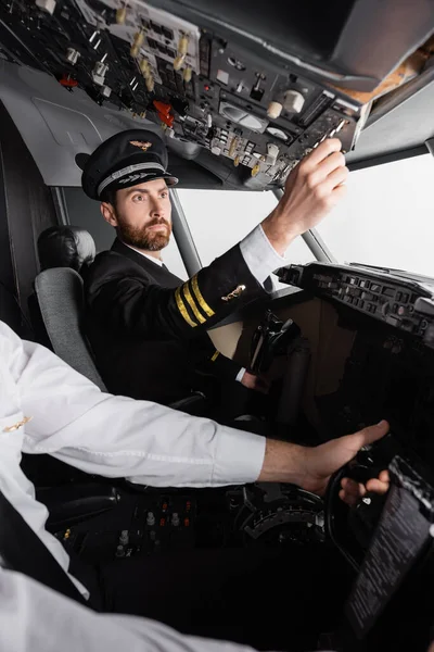 Piloto em cap e uniforme atingindo painel aéreo perto co-piloto usando jugo no simulador de avião — Fotografia de Stock