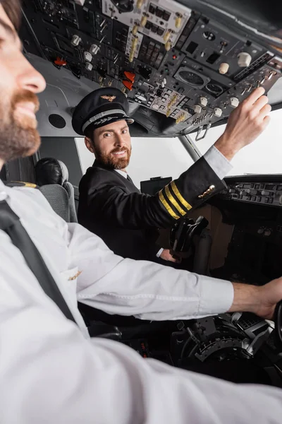Capitán alegre en el casquillo y uniforme alcanzando el panel superior cerca del copiloto en simulador de avión - foto de stock