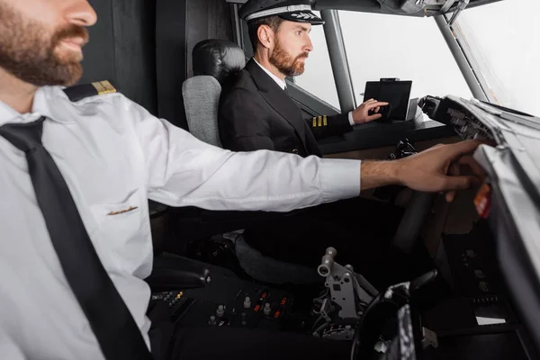 Kapitän in Mütze und Uniform mit Gerät mit leerem Bildschirm in der Nähe des Co-Piloten im Flugzeugsimulator — Stockfoto