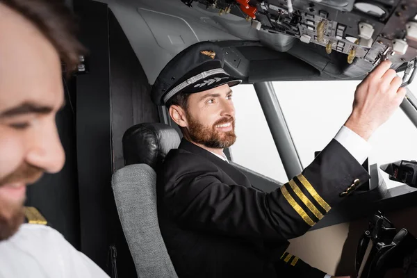 Piloto sonriente en uniforme alcanzando el panel superior cerca del copiloto borroso en simulador de avión - foto de stock