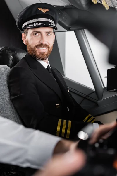 Pilota barbuto in uniforme e berretto sorridente vicino copilota sfocato — Foto stock