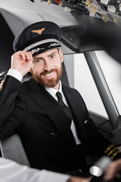 Bearded pilot in uniform adjusting cap and smiling near blurred co-pilot — Stock Photo