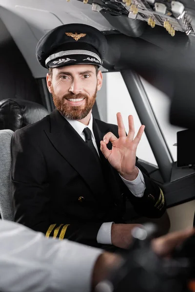 Piloto barbudo em uniforme mostrando ok e sorrindo perto de co-piloto turvo — Fotografia de Stock