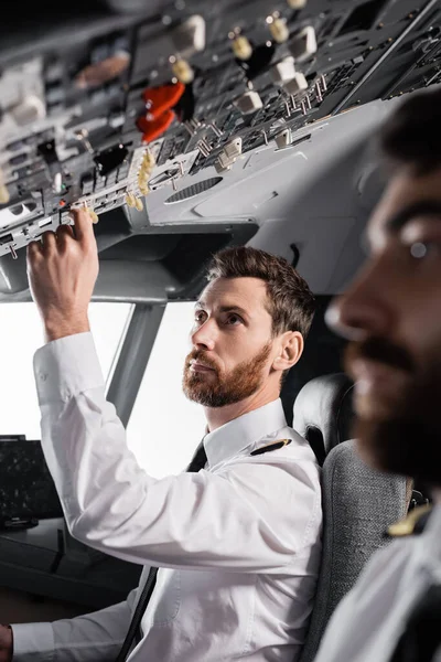 Piloto barbudo alcanzando el panel superior cerca del copiloto en simulador de avión - foto de stock