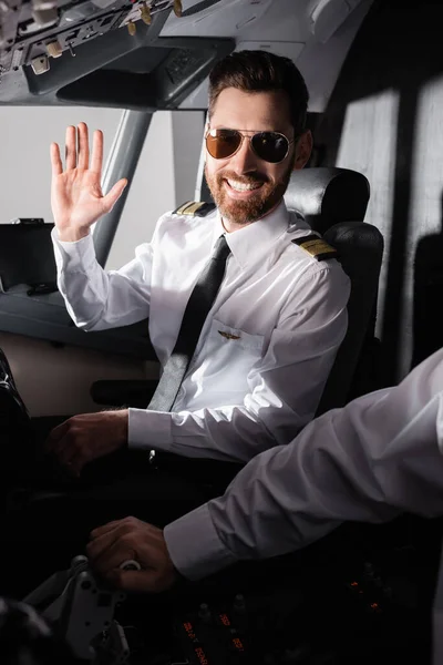 Fröhlicher Pilot mit Sonnenbrille winkt in der Nähe des Co-Piloten im Flugzeug — Stockfoto