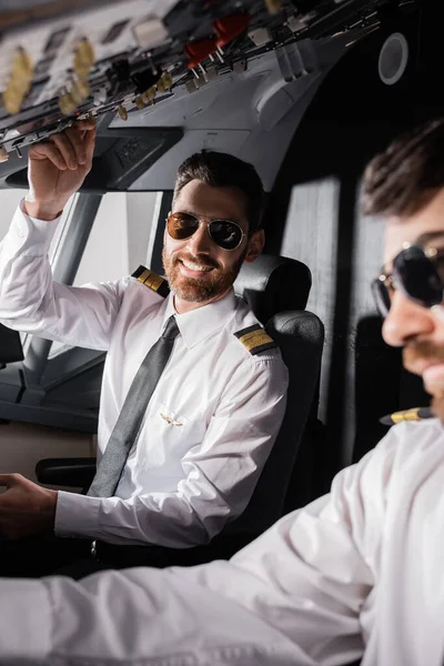 Piloto alegre en gafas de sol alcanzando el panel superior cerca del copiloto en simulador de avión - foto de stock