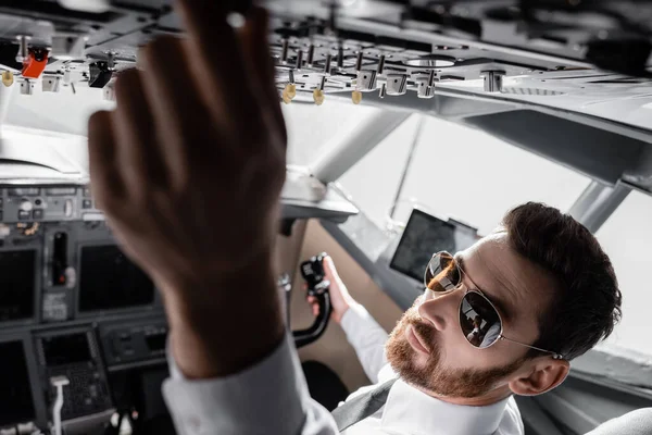 Vue grand angle du pilote barbu dans les lunettes de soleil atteignant le panneau supérieur dans le simulateur d'avion — Photo de stock