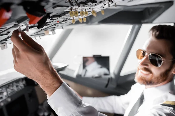 Piloto barbudo borroso en gafas de sol alcanzando el panel superior y sonriendo en simulador de avión - foto de stock