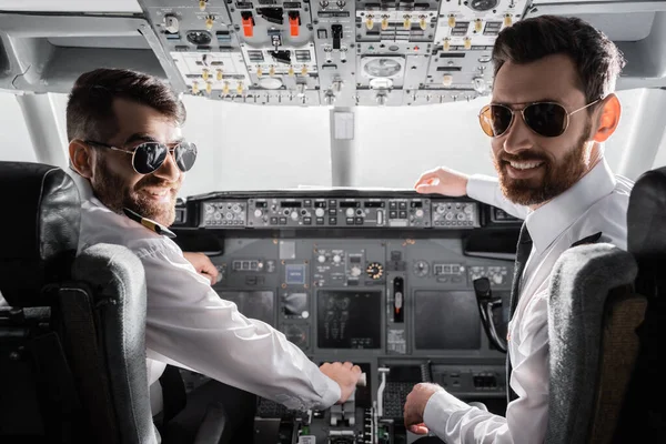 Happy pilots in sunglasses and uniform looking at camera in airplane simulator — Stock Photo