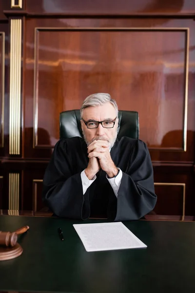 Juge aux cheveux gris en peignoir et lunettes assis les mains serrées près du procès sur le bureau — Photo de stock