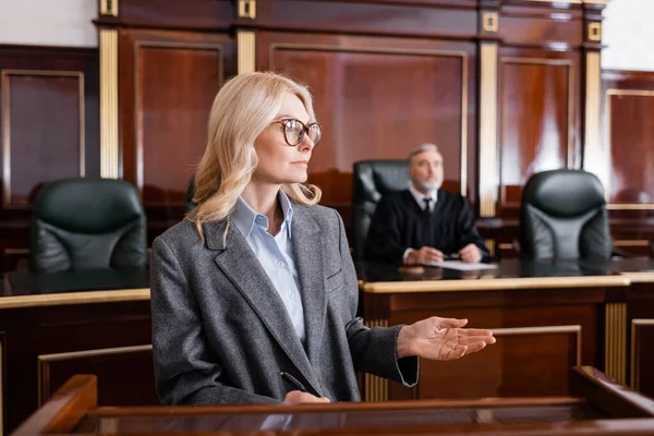 Blondine zeigt mit der Hand, während sie vor Gericht in der Nähe des Richters über Hintergründe spricht — Stockfoto