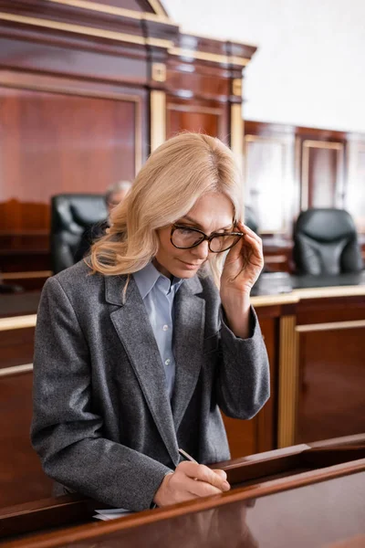 Blonde advocate adjusting eyeglasses while writing in courtroom — Stock Photo