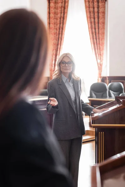 Blonde advocate pointing with hand while questioning blurred witness in court — Stock Photo