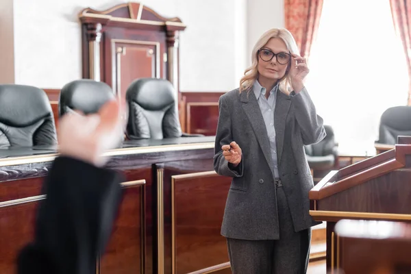 Avvocato maturo indicando con mano e aggiustando occhiali durante l'interrogatorio del testimone in tribunale — Foto stock