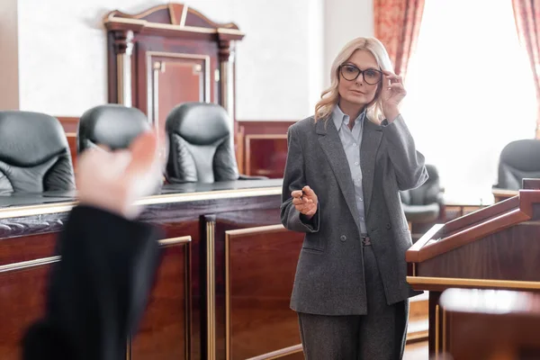 Abogada madura señalando con la mano y ajustando las gafas durante el interrogatorio de testigo en la corte — Stock Photo
