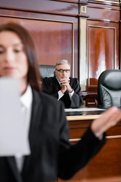 Fiscal borrosa hablando en la corte cerca del juez sentado con las manos apretadas en el fondo - foto de stock