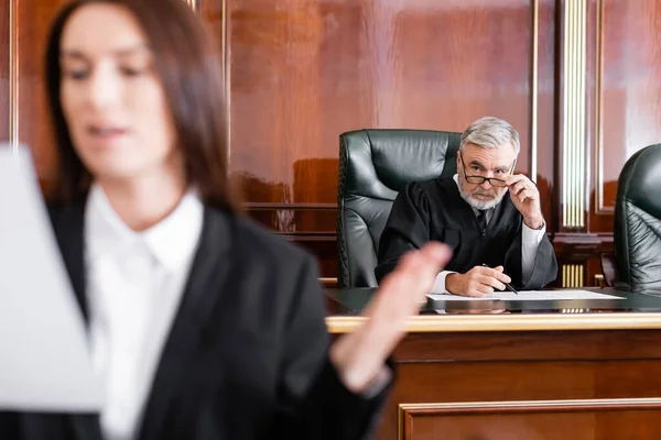 Grey-haired judge adjusting eyeglasses while looking at prosecutor speaking on blurred foreground — Stock Photo