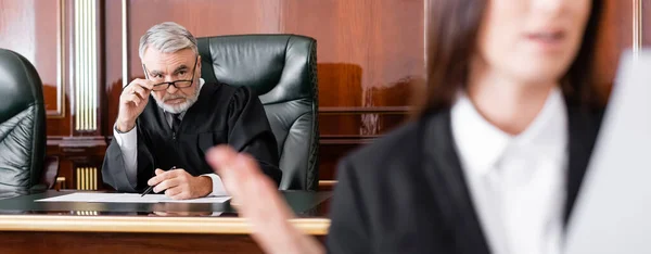 Senior judge adjusting eyeglasses while looking at prosecutor on blurred foreground, banner — Stock Photo