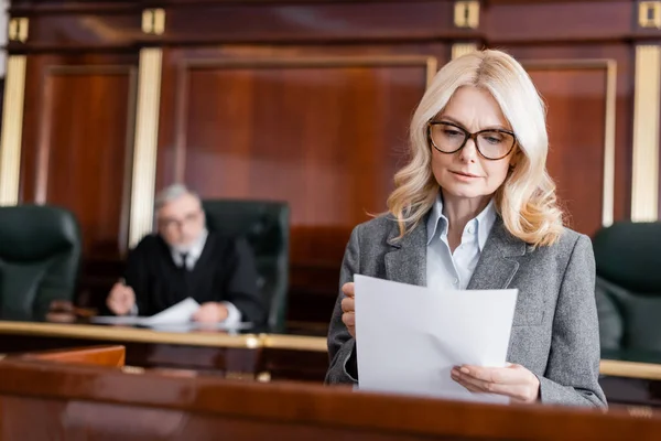Middle aged advocate in eyeglasses reading document while speaking in court — Stock Photo
