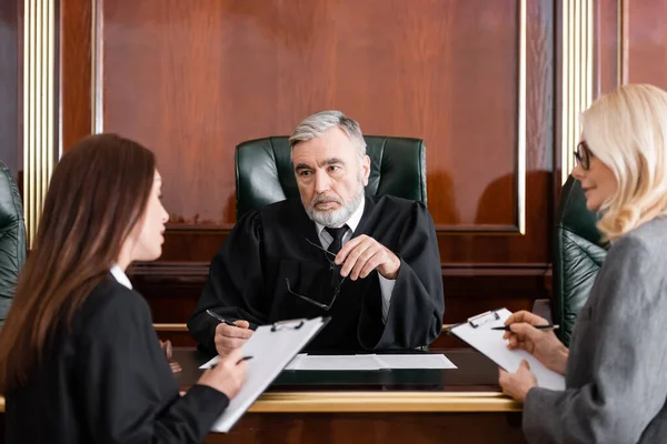 Senior judge holding eyeglasses near attorney and prosecutor writing on clipboards — Stock Photo