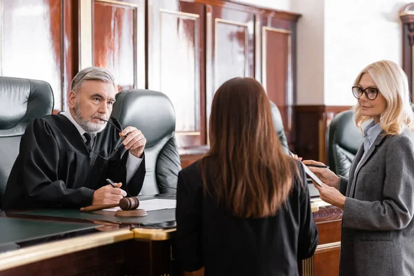 Thoughtful judge holding eyeglasses listen to prosecutor standing near attorney in court — Stock Photo