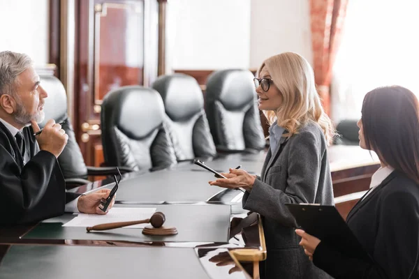 Blonde advocate with clipboard talking to senior judge while standing near prosecutor in court — Stock Photo