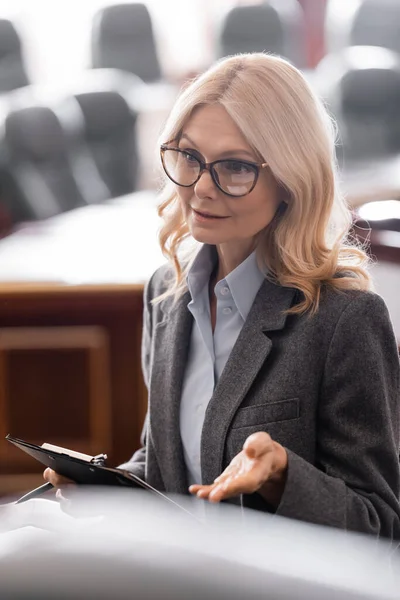Blonde middle aged advocate with clipboard talking in courtroom — Stock Photo