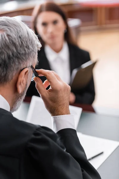 Grey-haired judge adjusting eyeglasses near blurred prosecutor in court — Stock Photo