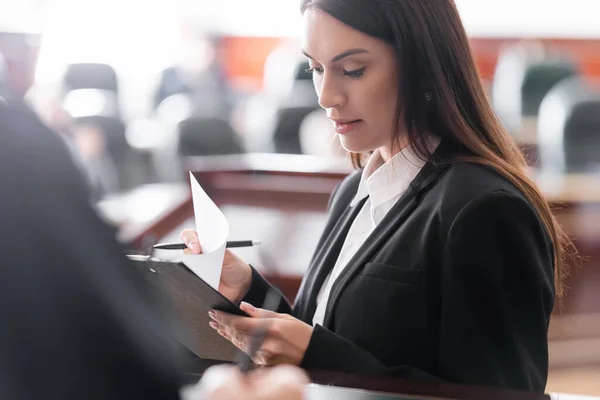 Brunette prosecutor reading lawsuit near blurred judge in courtroom — Stock Photo