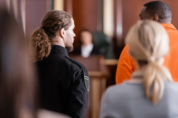 Vista trasera del hombre afroamericano acusado cerca de la guardia y los jurados borrosa en la sala del tribunal - foto de stock
