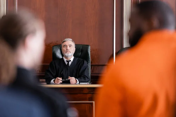Senior judge holding eyeglasses while looking at accused african american man on blurred foreground — Stock Photo