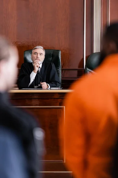 Thoughtful judge looking at blurred african american man standing near bailiff on blurred foreground — Stock Photo