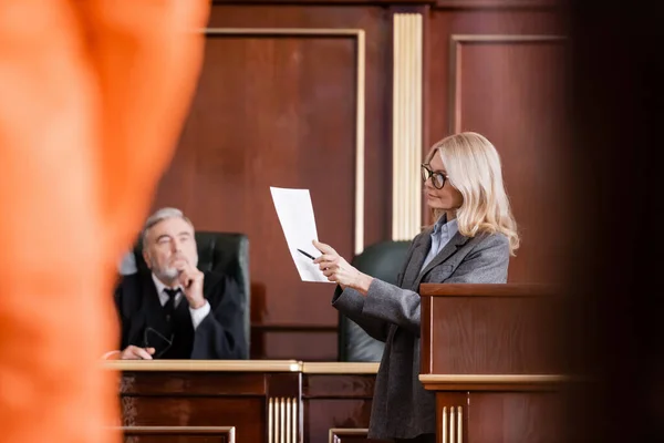 Blonde advocate pointing at lawsuit near judge in courtroom on blurred foreground — Stock Photo