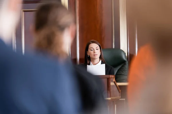 Fiscal leyendo demanda en el tribunal cerca de personas en primer plano borrosa - foto de stock