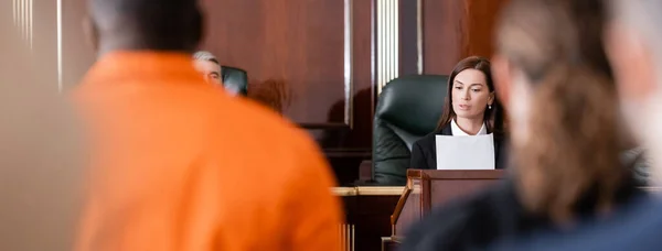 Fiscal leer documento cerca de juez y acusado hombre afroamericano en primer plano borrosa, bandera - foto de stock