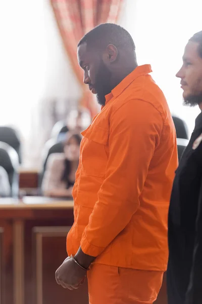 Vista lateral del hombre afroamericano esposado en uniforme de la cárcel cerca del alguacil y los jurados borrosos en la corte - foto de stock