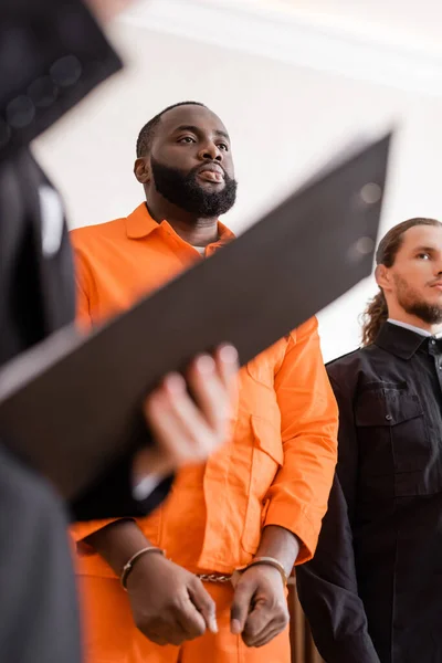 Acusado hombre afroamericano esposado de pie cerca del alguacil y fiscal borroso con portapapeles en la corte - foto de stock