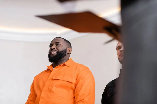 Vue en angle bas de l'homme afro-américain positif en uniforme de prison près de garde au tribunal sur le premier plan flou — Photo de stock