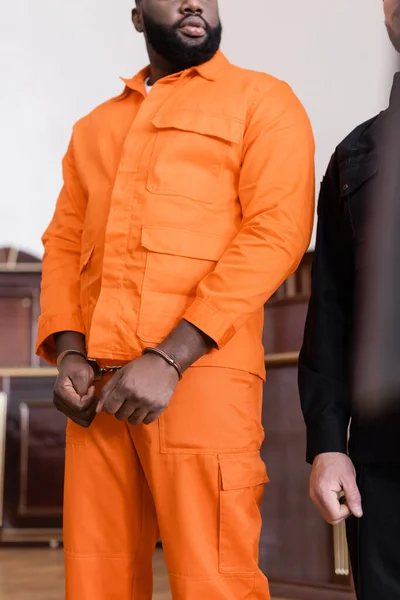 Visão parcial de homem afro-americano algemado em uniforme de prisão em pé perto de oficial de justiça em tribunal — Fotografia de Stock