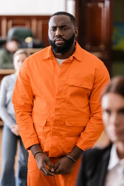 Frustrated african american man in handcuffs and jail uniform standing near blurred jurors in court — Stock Photo