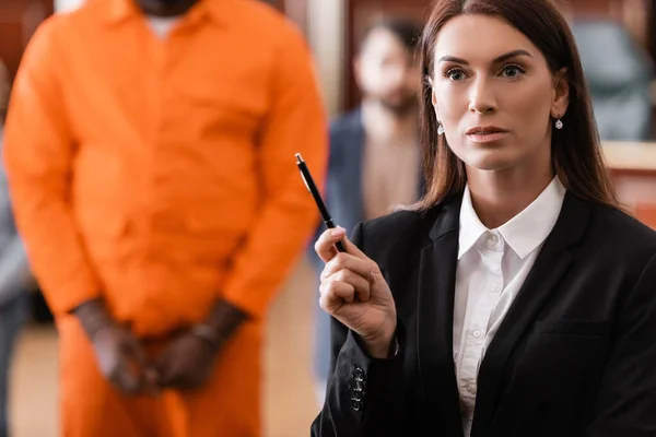 Brunette procureur en tenue formelle debout avec stylo près accusé homme afro-américain sur fond flou — Photo de stock