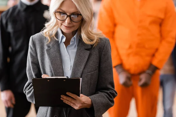 Attorney in eyeglasses writing on clipboard near accused african american man and guard on blurred background — Stock Photo