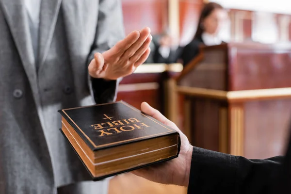 Vista recortada de los testigos jurando sobre la Biblia cerca de fiscal borrosa en la sala del tribunal - foto de stock