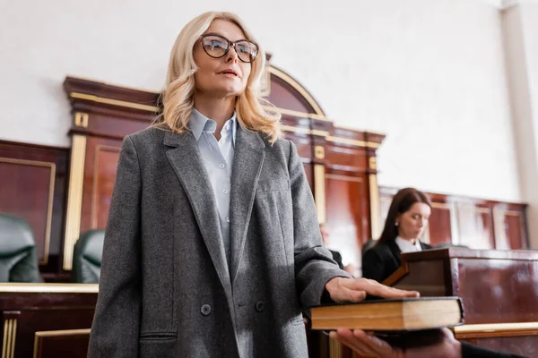 Femme dans les lunettes donnant jurer sur la bible au tribunal près du procureur sur fond flou — Photo de stock