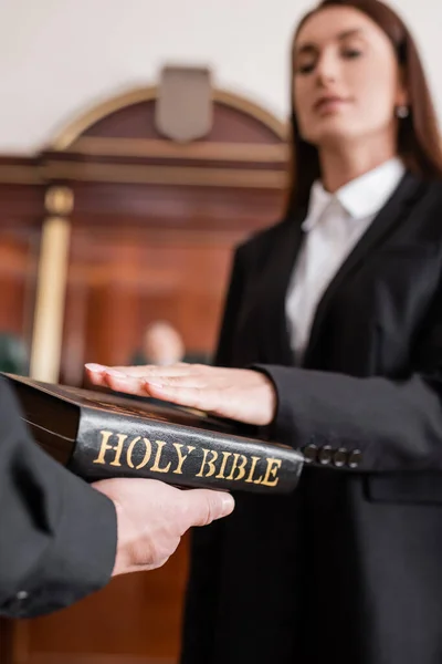 Blurred woman giving oath on bible in hand of bailiff in courtroom — Stock Photo