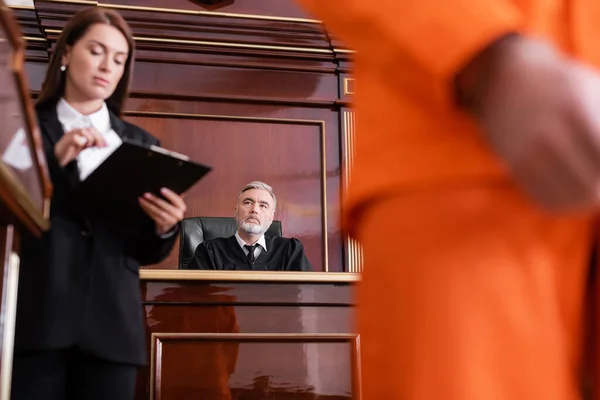 Prosecutor looking at clipboard near blurred man and senior judge on background — Stock Photo