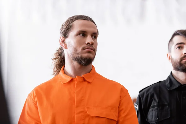 Accused man in orange uniform looking away near bailiff in court — Stock Photo