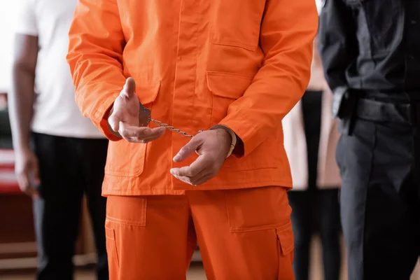 Partial view of handcuffed man in jail uniform near blurred guard and jurors in courtroom — Stock Photo