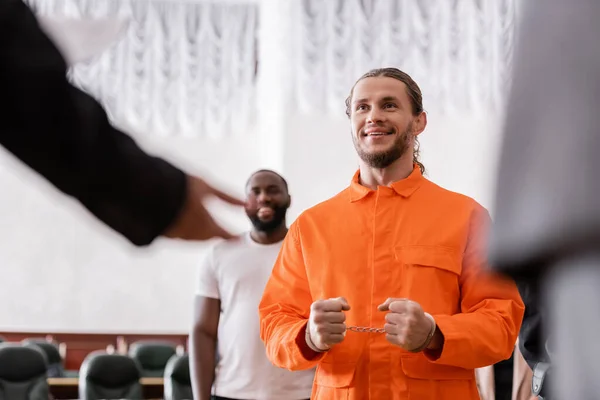 Happy man in handcuffs and jail uniform near blurred jurors in courtroom — Stock Photo